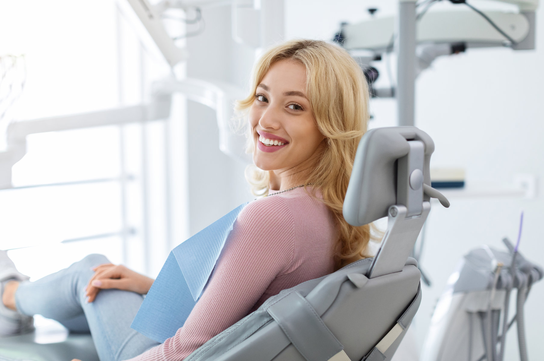 Smiling and relaxed young woman sitting at dental chair in modern clinic, waiting for her dentist. Happy attractive blonde lady attending luxury dental clinic, bacl view, copy space