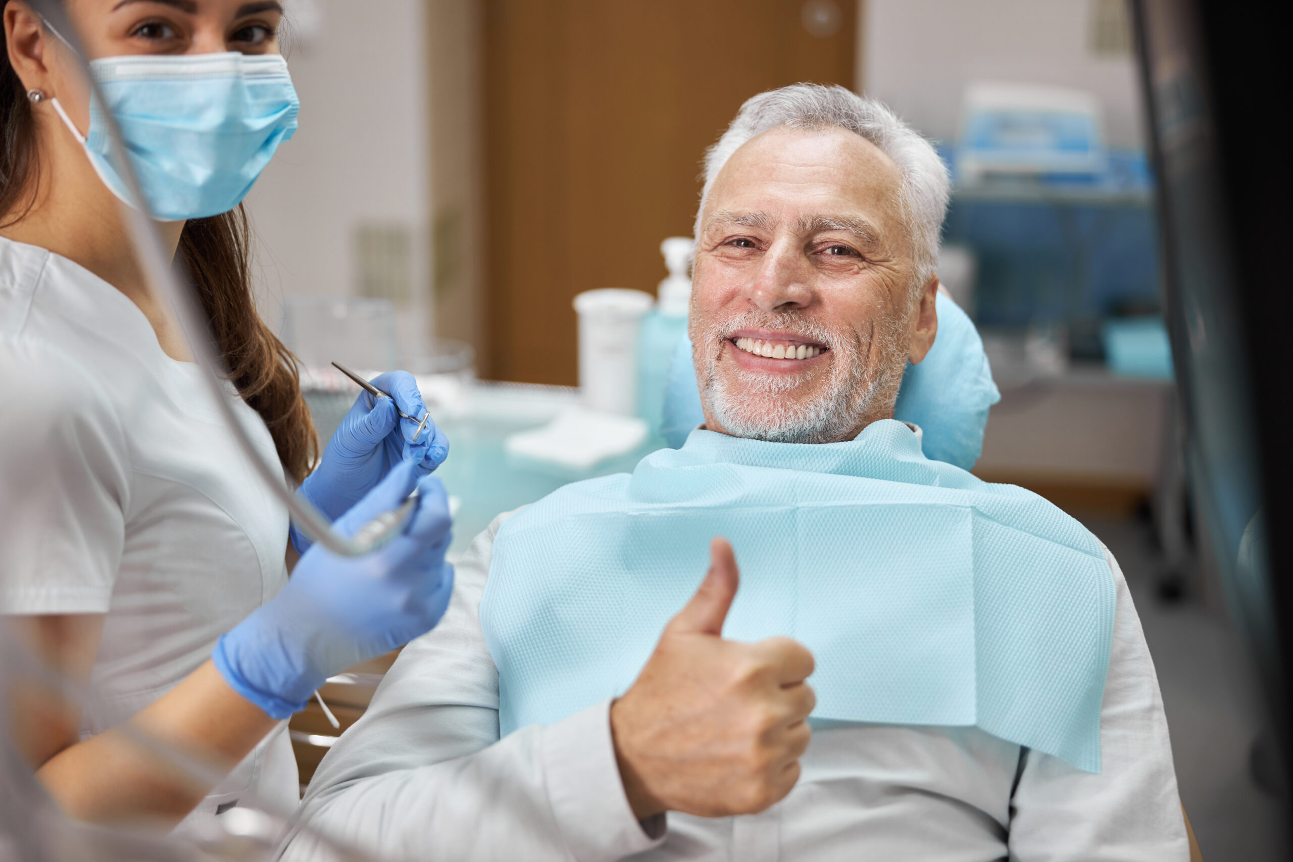 Mirthful senior citizen smiling and showing thumbs up at dental appointment with his dentist and looking at the camera